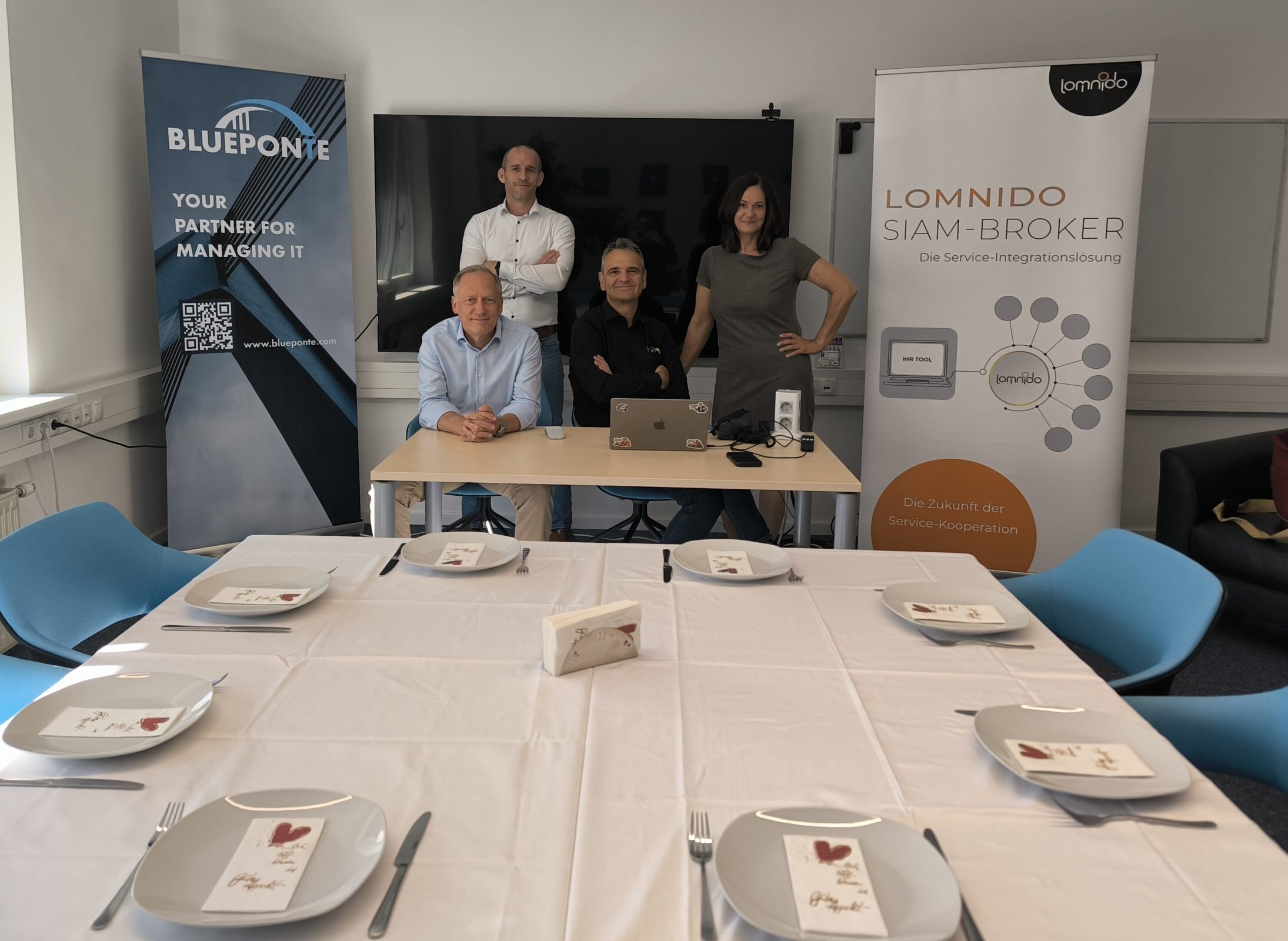 Business Breakfast von Blueponte und Lomnido mit Bannern der beidne Unternehmen. Vier Personen stehen und sitzen an einem Tisch in einem Meetingraum, davor ist ein gedeckter Tisch
